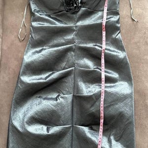 Puede incluir: Vestido plateado-gris, hasta la rodilla, con cuello en V y un solo tirante adornado con volantes y una flor de tela. El vestido mide aproximadamente 102 cm de largo, con una silueta ajustada y tejido texturizado.