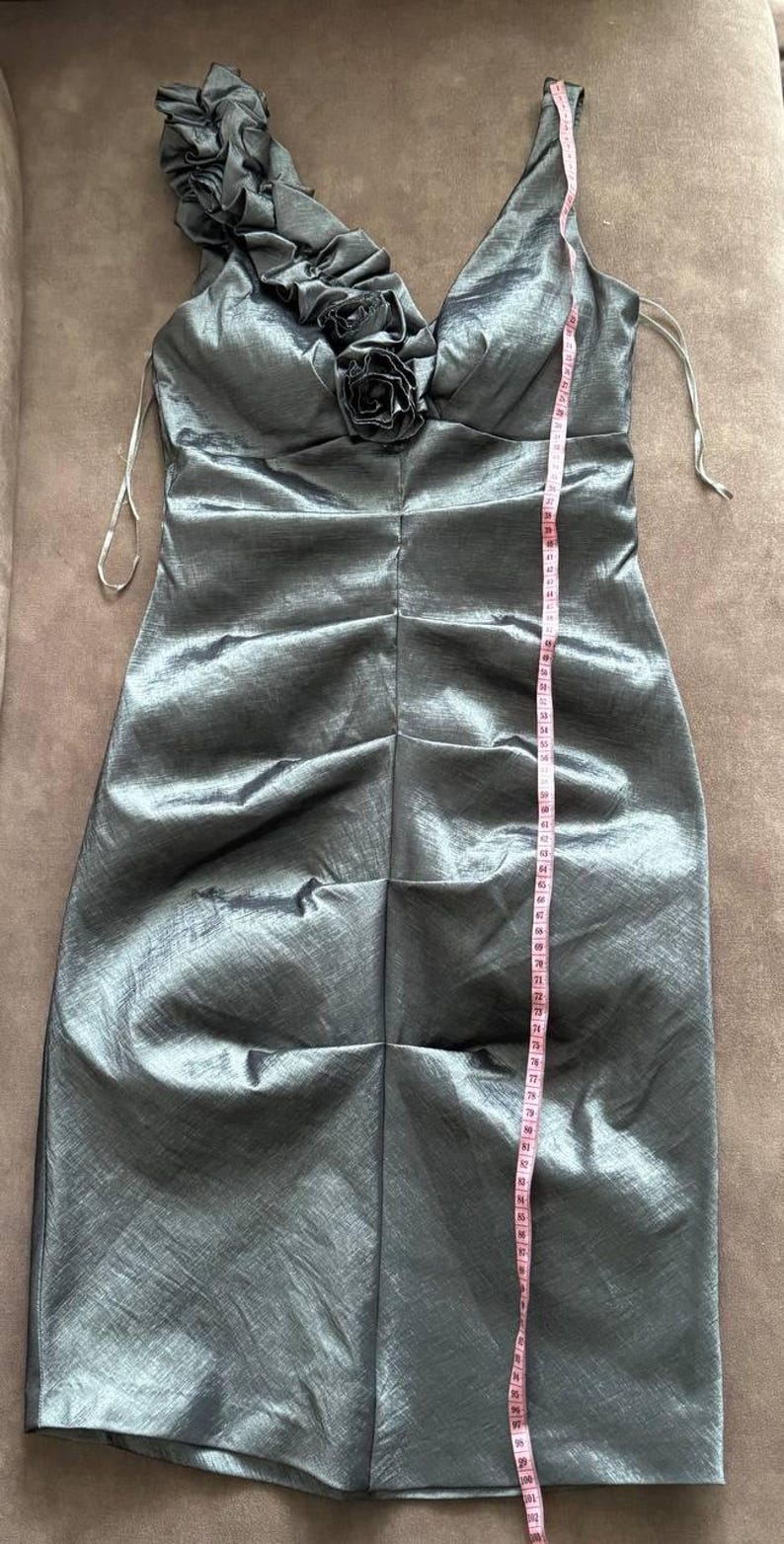 Puede incluir: Vestido plateado-gris, hasta la rodilla, con cuello en V y un solo tirante adornado con volantes y una flor de tela. El vestido mide aproximadamente 102 cm de largo, con una silueta ajustada y tejido texturizado.
