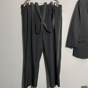 May include: Black suit pants with suspenders hanging on a hanger. The pants have a vertical stripe down the side. A black blazer is partially visible on the right side of the image.