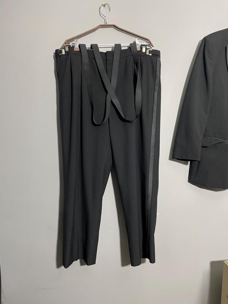 May include: Black suit pants with suspenders hanging on a hanger. The pants have a vertical stripe down the side. A black blazer is partially visible on the right side of the image.