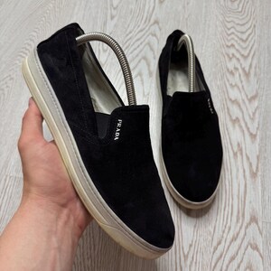 May include: Black suede slip-on shoes with a white sole. The shoes have the brand name "Prada" on the side. The shoes are designed for casual wear.