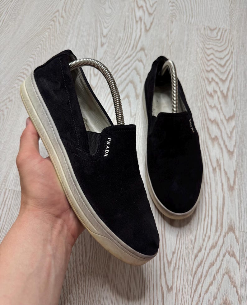 May include: Black suede slip-on shoes with a white sole. The shoes have the brand name "Prada" on the side. The shoes are designed for casual wear.