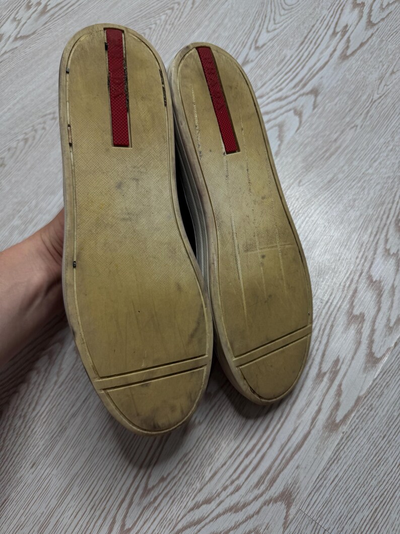 May include: A pair of worn sneakers with off-white soles and a red accent on the heel. The soles are dirty, showing signs of wear and use. The shoes are likely casual footwear.