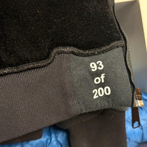 Pode incluir: Close-up de um moletom preto com textura aveludada na parte superior. O n&uacute;mero "93 of 200" est&aacute; impresso em branco em uma faixa preta perto do punho. Um z&iacute;per &eacute; vis&iacute;vel na lateral.