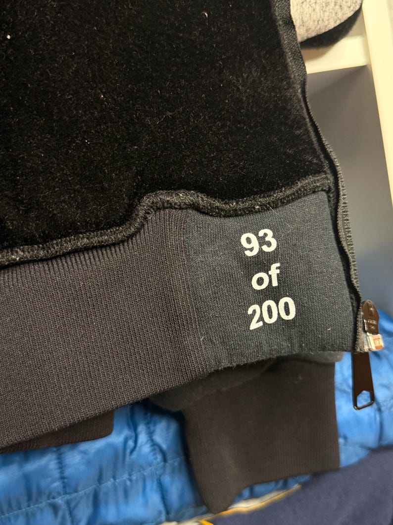 Pode incluir: Close-up de um moletom preto com textura aveludada na parte superior. O n&uacute;mero "93 of 200" est&aacute; impresso em branco em uma faixa preta perto do punho. Um z&iacute;per &eacute; vis&iacute;vel na lateral.