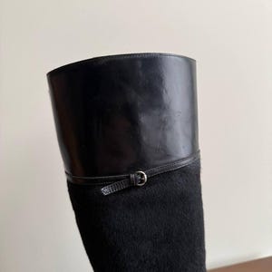 Op de afbeelding: Zwarte rijlaars met een leren bovenwerk en een getextureerde, vachtachtige schacht. De laars heeft een leren band met een zilverkleurige gesp. Het ontwerp van de laars is klassiek en elegant.
