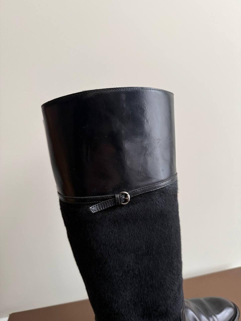 Op de afbeelding: Zwarte rijlaars met een leren bovenwerk en een getextureerde, vachtachtige schacht. De laars heeft een leren band met een zilverkleurige gesp. Het ontwerp van de laars is klassiek en elegant.
