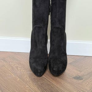 May include: A pair of black suede knee-high boots. The boots have a rounded toe and a block heel. The boots are standing on a wooden floor. The boots have a zipper on the side.