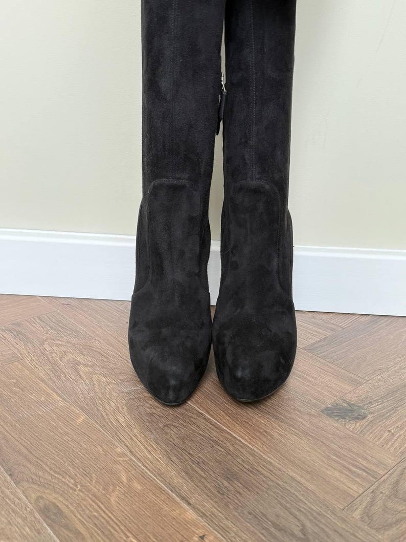 May include: A pair of black suede knee-high boots. The boots have a rounded toe and a block heel. The boots are standing on a wooden floor. The boots have a zipper on the side.