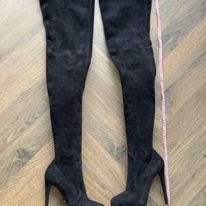 May include: A pair of black suede over-the-knee boots with stiletto heels. The boots have a platform sole and a tall shaft that extends above the knee. A pink measuring tape is placed next to the boots.