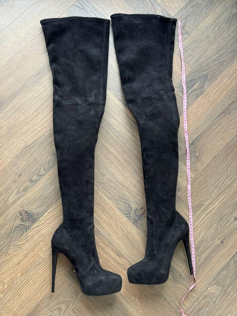 May include: A pair of black suede over-the-knee boots with stiletto heels. The boots have a platform sole and a tall shaft that extends above the knee. A pink measuring tape is placed next to the boots.