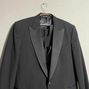 May include: A formal black tuxedo jacket with a satin lapel, hanging on a metal hanger. The jacket has a single-button closure and a chest pocket. The image shows the front of the jacket, with a glimpse of the matching vest underneath.