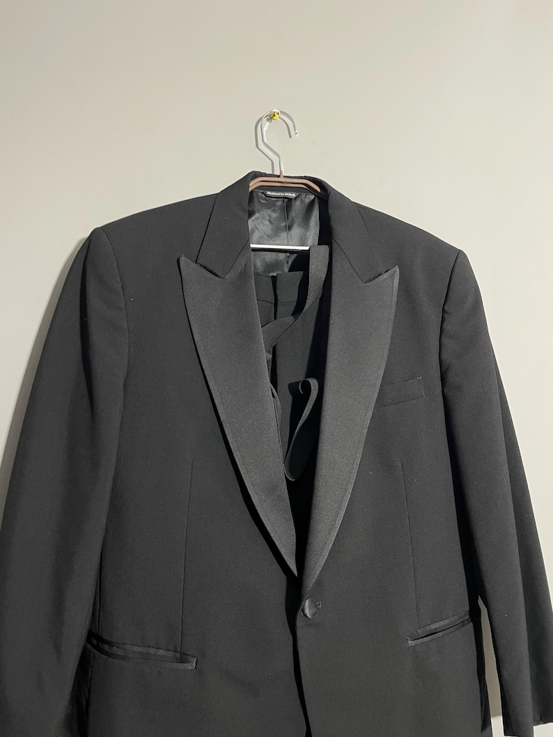 May include: A formal black tuxedo jacket with a satin lapel, hanging on a metal hanger. The jacket has a single-button closure and a chest pocket. The image shows the front of the jacket, with a glimpse of the matching vest underneath.