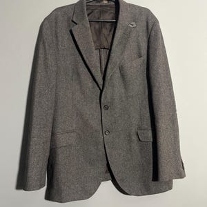 Op de afbeelding: Een grijs tweed blazer met een drieknoopsluiting en een klein speldje op de revers.