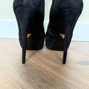 May include: A pair of black suede high-heeled boots. The boots have a pointed toe and a stiletto heel. The heels are approximately 5 inches tall. The soles of the boots have a gold logo.