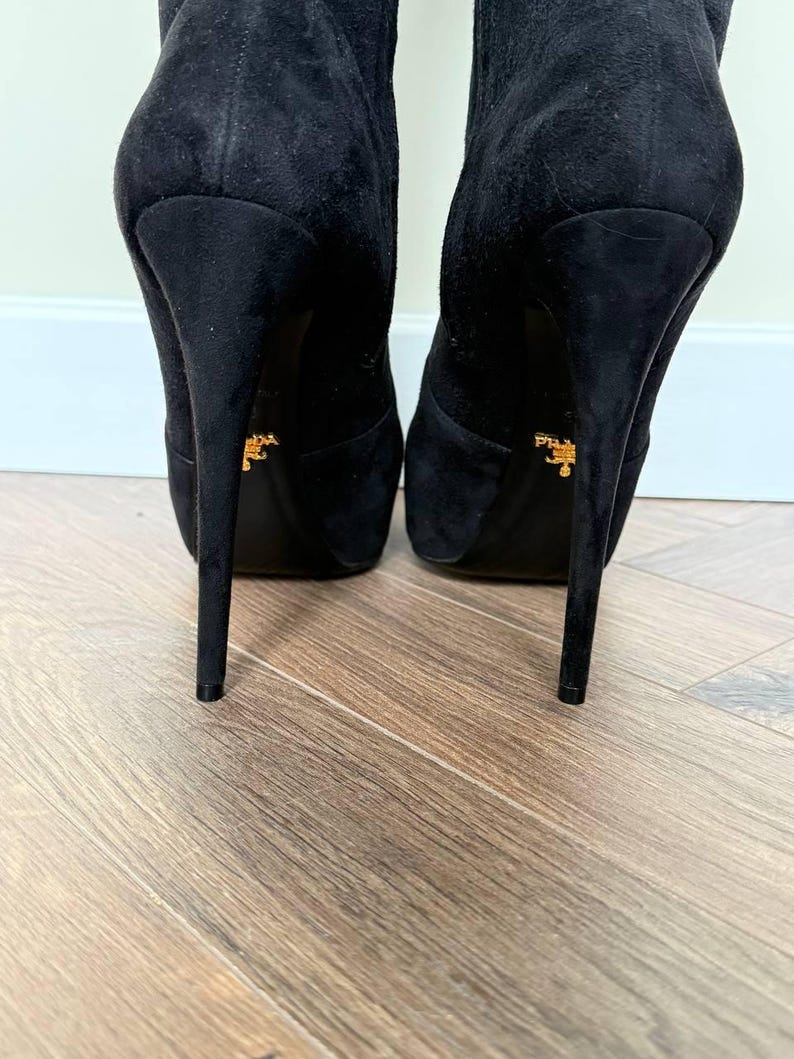 May include: A pair of black suede high-heeled boots. The boots have a pointed toe and a stiletto heel. The heels are approximately 5 inches tall. The soles of the boots have a gold logo.