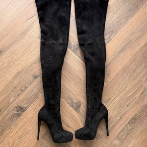 May include: A pair of black suede over-the-knee boots with stiletto heels and platform soles. The boots are displayed on a wooden surface, showcasing their sleek design and high fashion appeal.