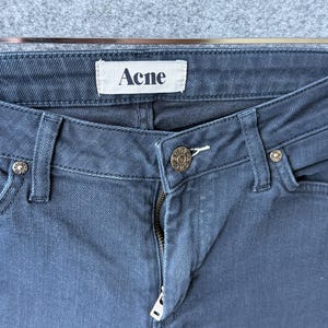 Op de afbeelding: Donkerblauwe spijkerbroek van denim met een wit binnenlabel met de tekst Acne in zwarte letters. De broek is voorzien van een metalen knoop met de inscriptie Acne, een metalen ritssluiting en zilverkleurige klinknagels op de zakken.
