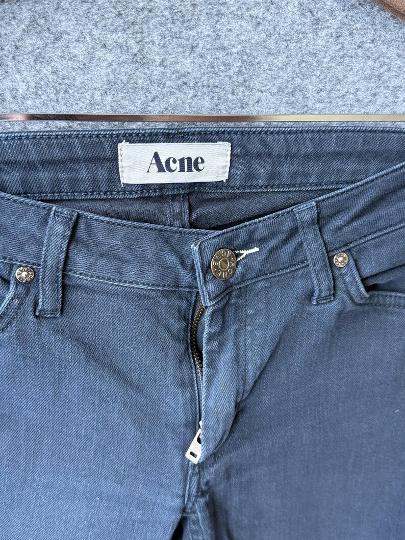 Op de afbeelding: Donkerblauwe spijkerbroek van denim met een wit binnenlabel met de tekst Acne in zwarte letters. De broek is voorzien van een metalen knoop met de inscriptie Acne, een metalen ritssluiting en zilverkleurige klinknagels op de zakken.