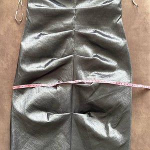 Puede incluir: Un vestido plateado-gris sin tirantes con un corpi&ntilde;o fruncido y una flor decorativa en el busto. El vestido se mide con una cinta m&eacute;trica rosa. El vestido parece estar hecho de una tela brillante y fluida.