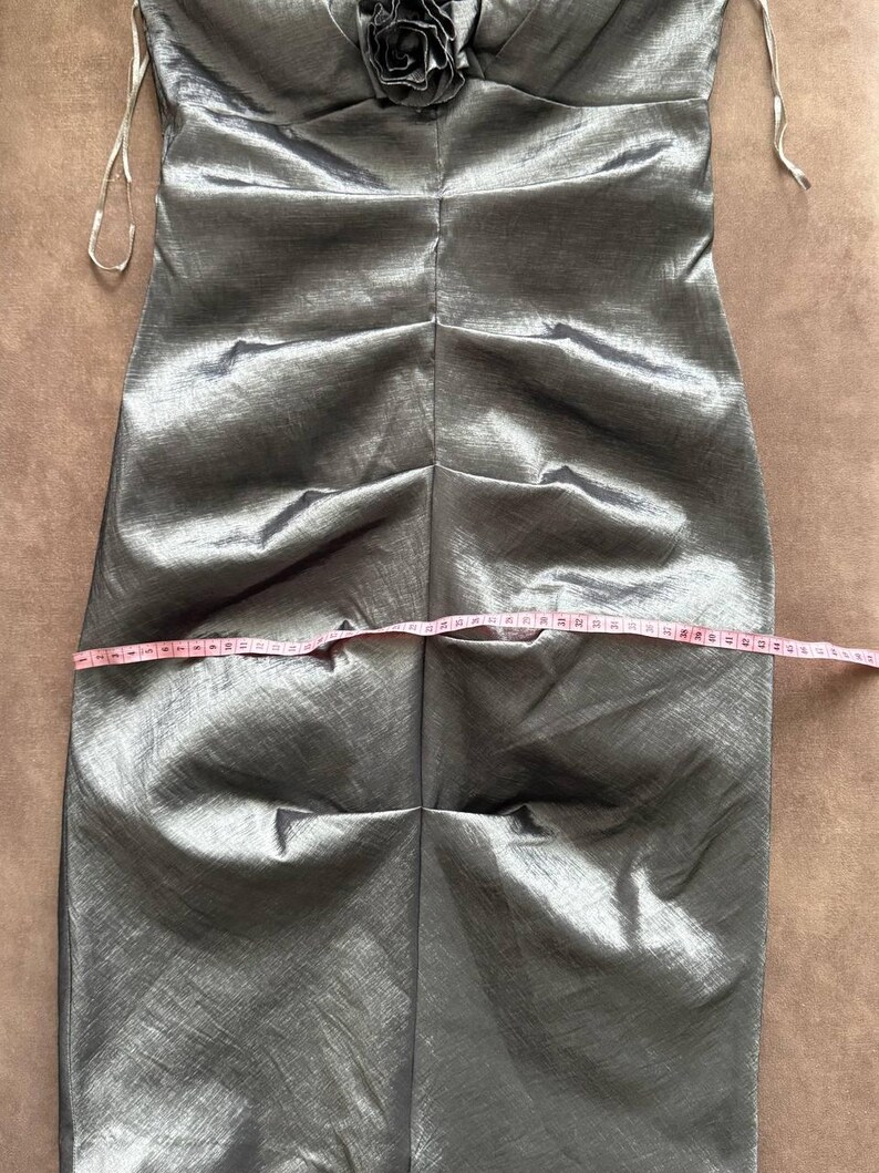 Puede incluir: Un vestido plateado-gris sin tirantes con un corpi&ntilde;o fruncido y una flor decorativa en el busto. El vestido se mide con una cinta m&eacute;trica rosa. El vestido parece estar hecho de una tela brillante y fluida.