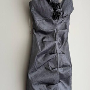 Puede incluir: Un vestido gris de un solo hombro con un escote con volantes y detalles florales. El vestido est&aacute; colgado de una percha de madera. La tela tiene un aspecto texturizado y el vestido est&aacute; fruncido en la cintura.