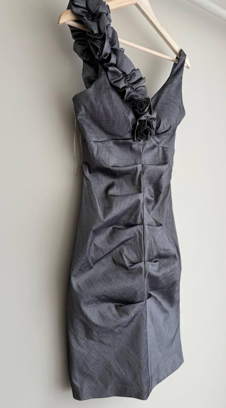 Puede incluir: Un vestido gris de un solo hombro con un escote con volantes y detalles florales. El vestido est&aacute; colgado de una percha de madera. La tela tiene un aspecto texturizado y el vestido est&aacute; fruncido en la cintura.