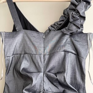 Puede incluir: Un vestido gris sin mangas con un brillo met&aacute;lico texturizado. El vestido presenta un solo tirante con volantes y una cremallera en la espalda. El vestido est&aacute; colgado de una percha de madera.
