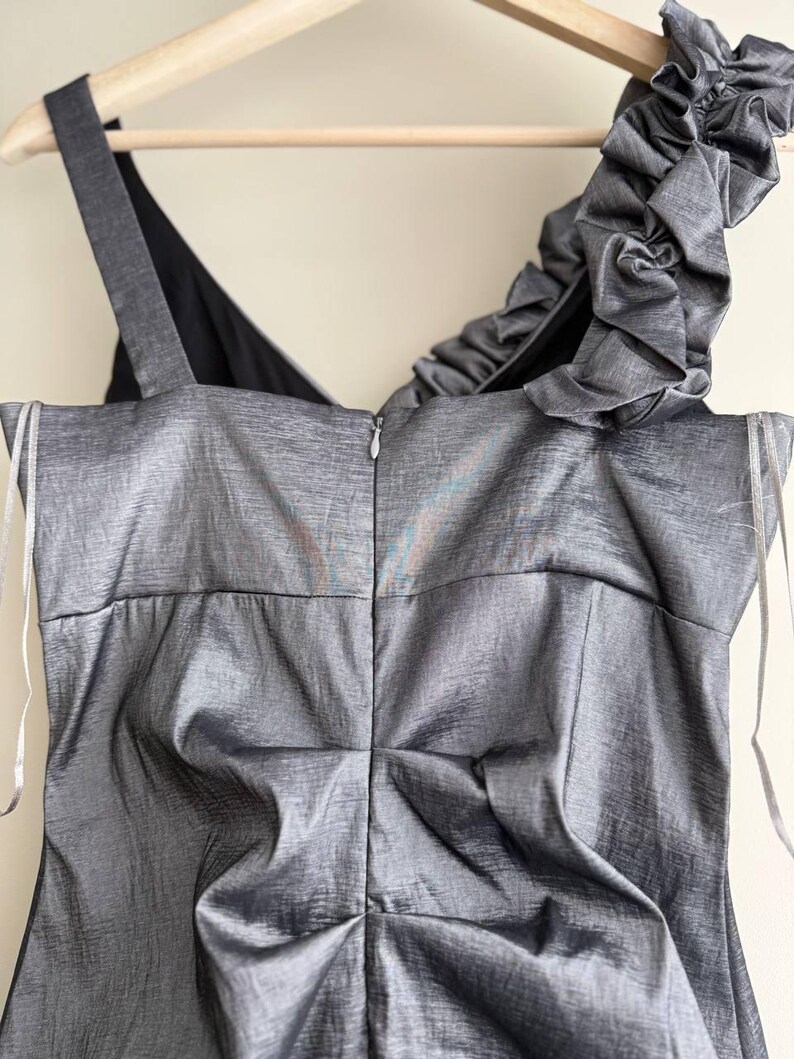 Puede incluir: Un vestido gris sin mangas con un brillo met&aacute;lico texturizado. El vestido presenta un solo tirante con volantes y una cremallera en la espalda. El vestido est&aacute; colgado de una percha de madera.