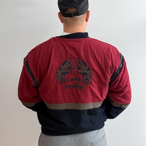 May include: A red, black, and brown yachting jacket with the text "Paul & Shark Yachting" on the back. The jacket has a black collar and black accents on the sleeves. A black baseball cap is worn.