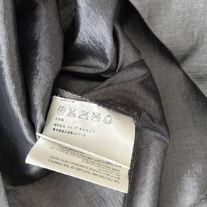 Puede incluir: Primer plano de una tela gris oscuro y brillante. Una etiqueta blanca con texto japon&eacute;s y s&iacute;mbolos de cuidado est&aacute; adjunta. La tela tiene un brillo sutil, lo que sugiere un material ligero, posiblemente seda o una mezcla.