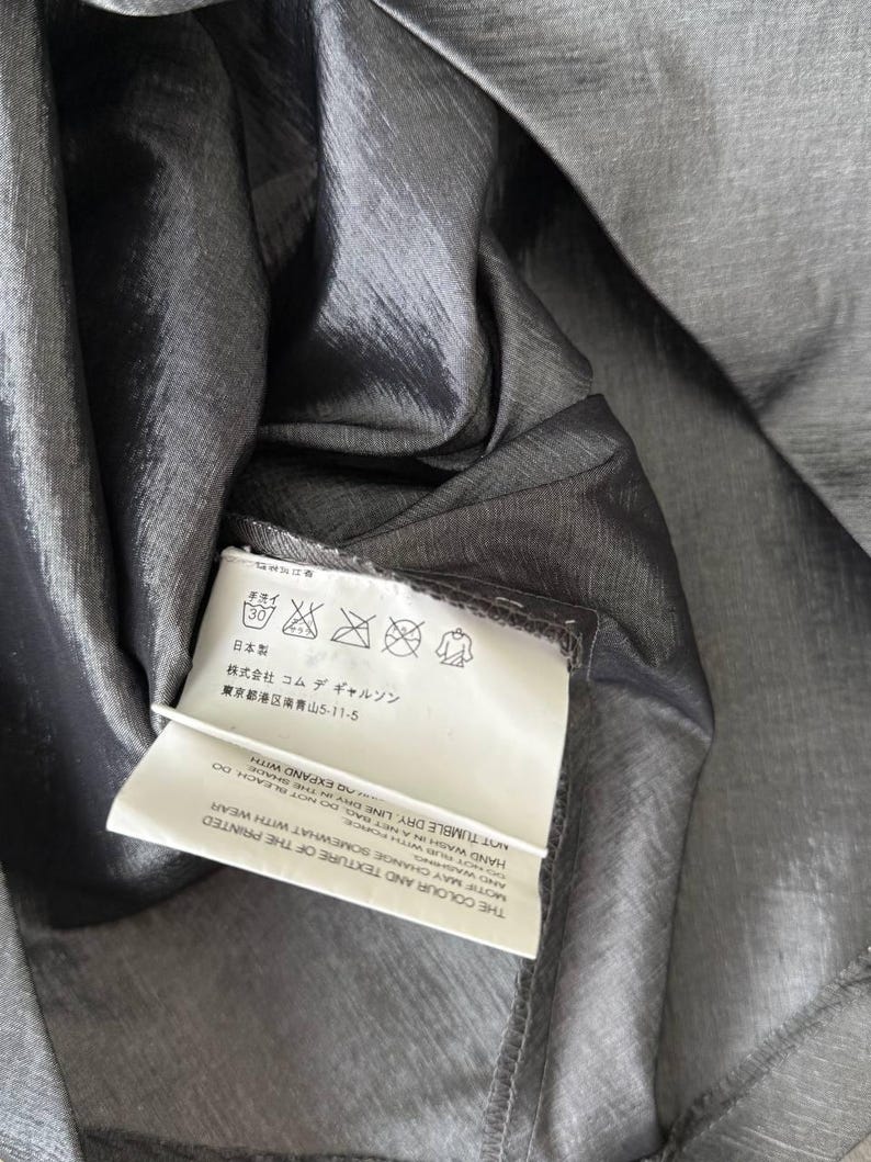 Puede incluir: Primer plano de una tela gris oscuro y brillante. Una etiqueta blanca con texto japon&eacute;s y s&iacute;mbolos de cuidado est&aacute; adjunta. La tela tiene un brillo sutil, lo que sugiere un material ligero, posiblemente seda o una mezcla.