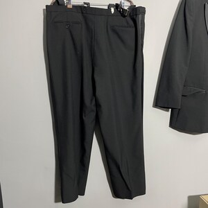 May include: Black suit pants hanging on a metal hanger, with a matching black blazer to the right. The pants have suspender buttons and a single button closure. The blazer is partially visible.