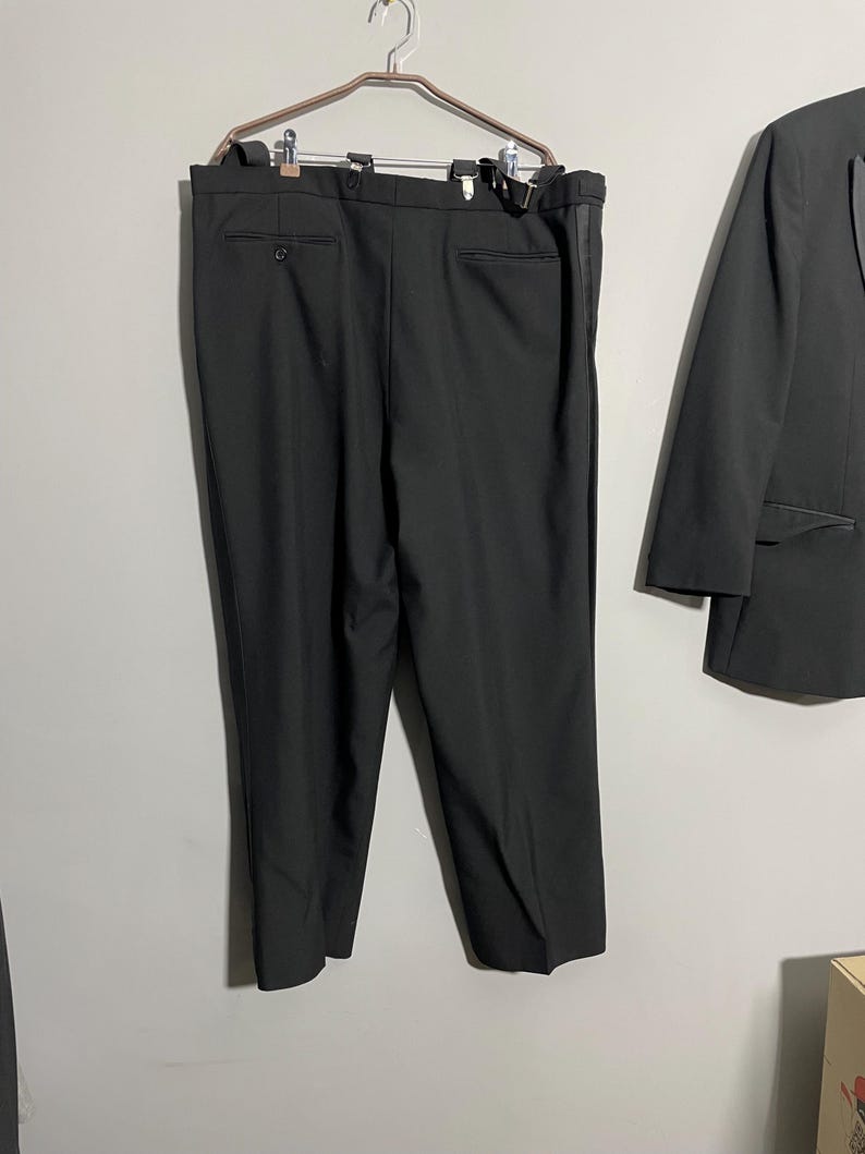 May include: Black suit pants hanging on a metal hanger, with a matching black blazer to the right. The pants have suspender buttons and a single button closure. The blazer is partially visible.