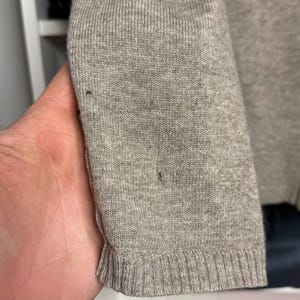 Pu&ograve; includere: Primo piano di una manica di maglione grigio chiaro. La manica &egrave; realizzata in tessuto a maglia con polsino a coste. Il maglione ha un design semplice e classico, adatto all'abbigliamento casual. Il tessuto appare morbido e confortevole.
