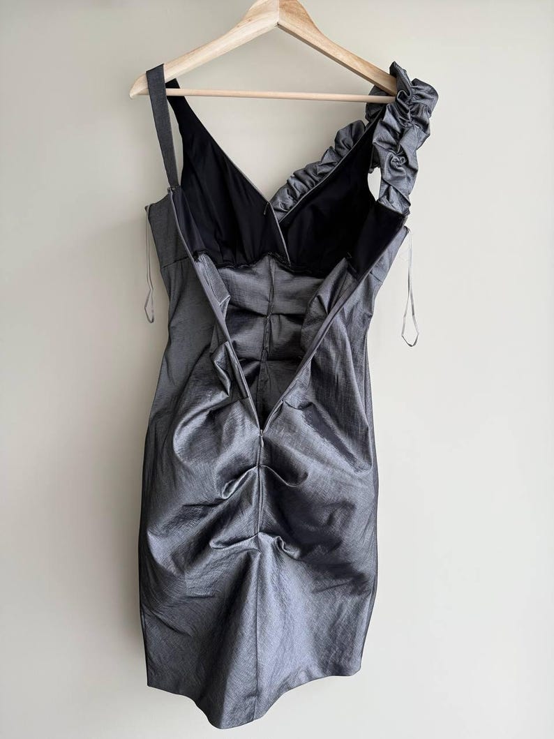 Puede incluir: Un vestido gris sin mangas con cuello en V y una sola correa. El vestido tiene un dise&ntilde;o fruncido en el corpi&ntilde;o y el hombro, y est&aacute; colgado de una percha de madera. El vestido est&aacute; hecho de una tela brillante.