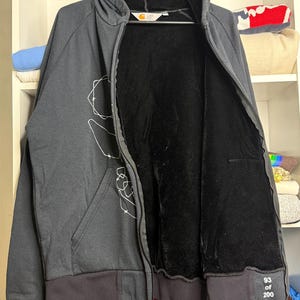 Pode incluir: Um casaco com capuz preto com z&iacute;per e forro interno preto. O casaco tem um gr&aacute;fico branco na frente e bainha e punhos canelados pretos. O texto "93 of 200" est&aacute; impresso no punho. O logotipo da Carhartt &eacute; vis&iacute;vel.