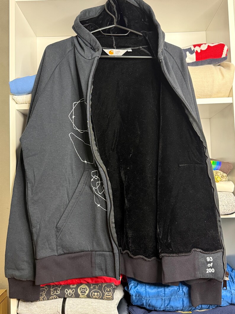 Pode incluir: Um casaco com capuz preto com z&iacute;per e forro interno preto. O casaco tem um gr&aacute;fico branco na frente e bainha e punhos canelados pretos. O texto "93 of 200" est&aacute; impresso no punho. O logotipo da Carhartt &eacute; vis&iacute;vel.