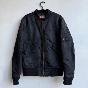 May include: Black bomber jacket with a ribbed collar, cuffs, and hem. Features a zip-up front, multiple pockets, and a small logo patch on the chest. The jacket is hanging on a black hanger against a white wall.