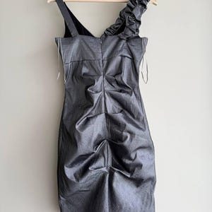 Puede incluir: Un vestido gris, hasta la rodilla, con un corpi&ntilde;o fruncido y una sola correa para el hombro. El vestido presenta un detalle con volantes en el hombro y est&aacute; colgado de una percha de madera. La tela tiene un brillo sutil.