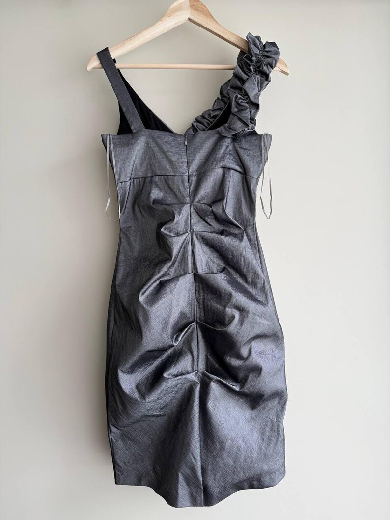 Puede incluir: Un vestido gris, hasta la rodilla, con un corpi&ntilde;o fruncido y una sola correa para el hombro. El vestido presenta un detalle con volantes en el hombro y est&aacute; colgado de una percha de madera. La tela tiene un brillo sutil.