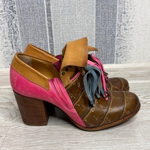 May include: A pair of unique ankle boots with a mix of textures and colors. The boots feature brown, pink, and tan leather, with a block heel and decorative fringe. The toe area has a crocodile skin pattern.
