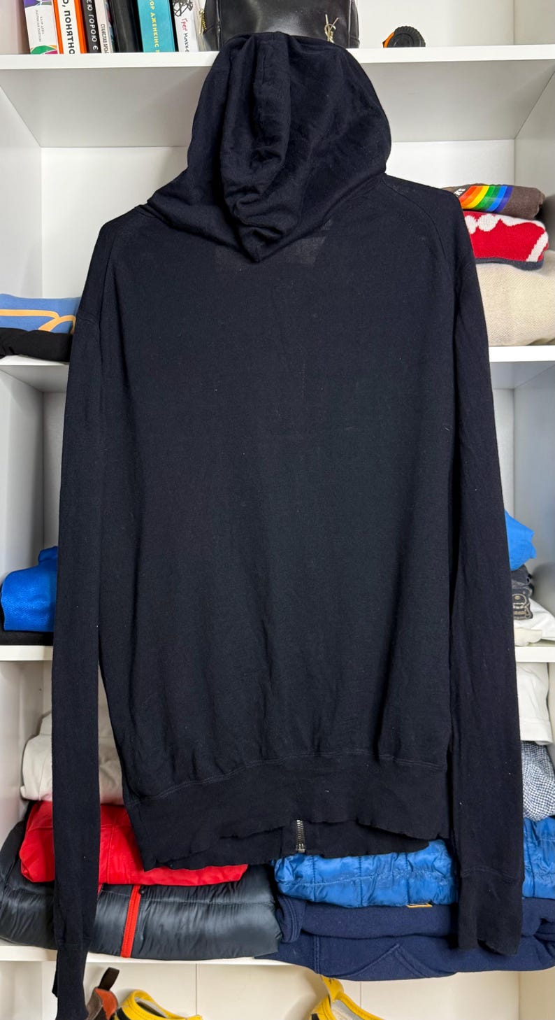 May include: Black hooded sweatshirt with a full-length zipper. The sweatshirt is made of a soft material and features a hood and long sleeves. The garment is displayed on a shelf with other clothing items.