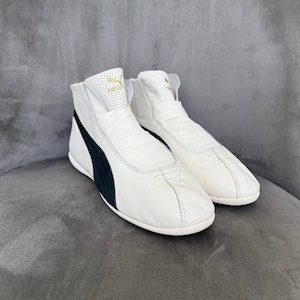 May include: Pair of white Puma high-top sneakers with a black stripe on the side. The sneakers have a perforated tongue and the Puma logo in gold. The shoes are made of leather and have a classic, sporty design.