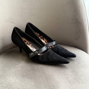 May include: A pair of black suede pumps with pointed toes and kitten heels. The shoes feature a strap across the top with two decorative buttons and a leopard print interior. The shoes are displayed on a neutral-toned chair.