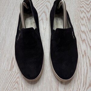May include: A pair of black suede slip-on shoes with white soles. The shoes have the brand name "PRADA" in white lettering on the side. Shoe trees are inserted in the shoes.