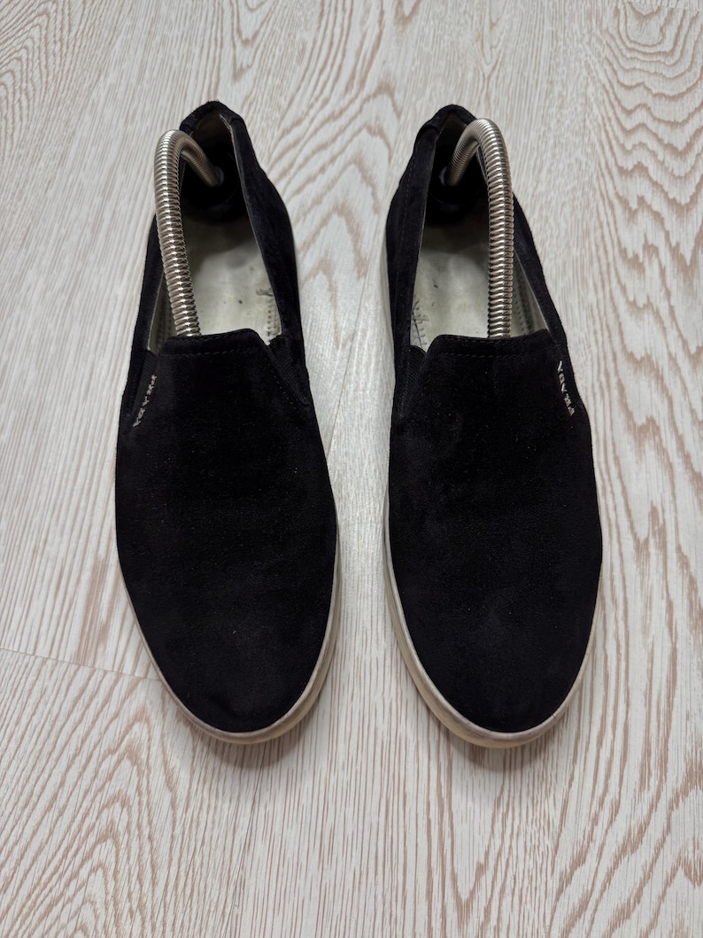 May include: A pair of black suede slip-on shoes with white soles. The shoes have the brand name "PRADA" in white lettering on the side. Shoe trees are inserted in the shoes.