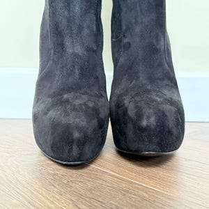 May include: Close-up of two black suede boots. The boots have a rounded toe and a block heel. The suede material has a soft, textured appearance. The boots are set against a light-colored wall and a wooden floor.