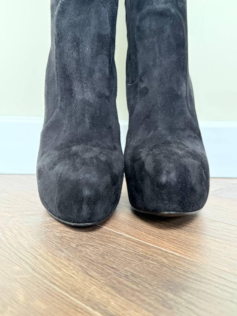May include: Close-up of two black suede boots. The boots have a rounded toe and a block heel. The suede material has a soft, textured appearance. The boots are set against a light-colored wall and a wooden floor.
