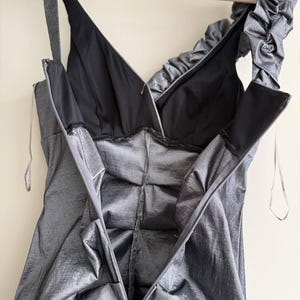 Puede incluir: Un vestido gris plateado con cuello en V y forro negro, colgado de una percha de madera. El vestido presenta una cremallera en la espalda y un detalle de volantes en el hombro. La tela tiene un brillo sutil.